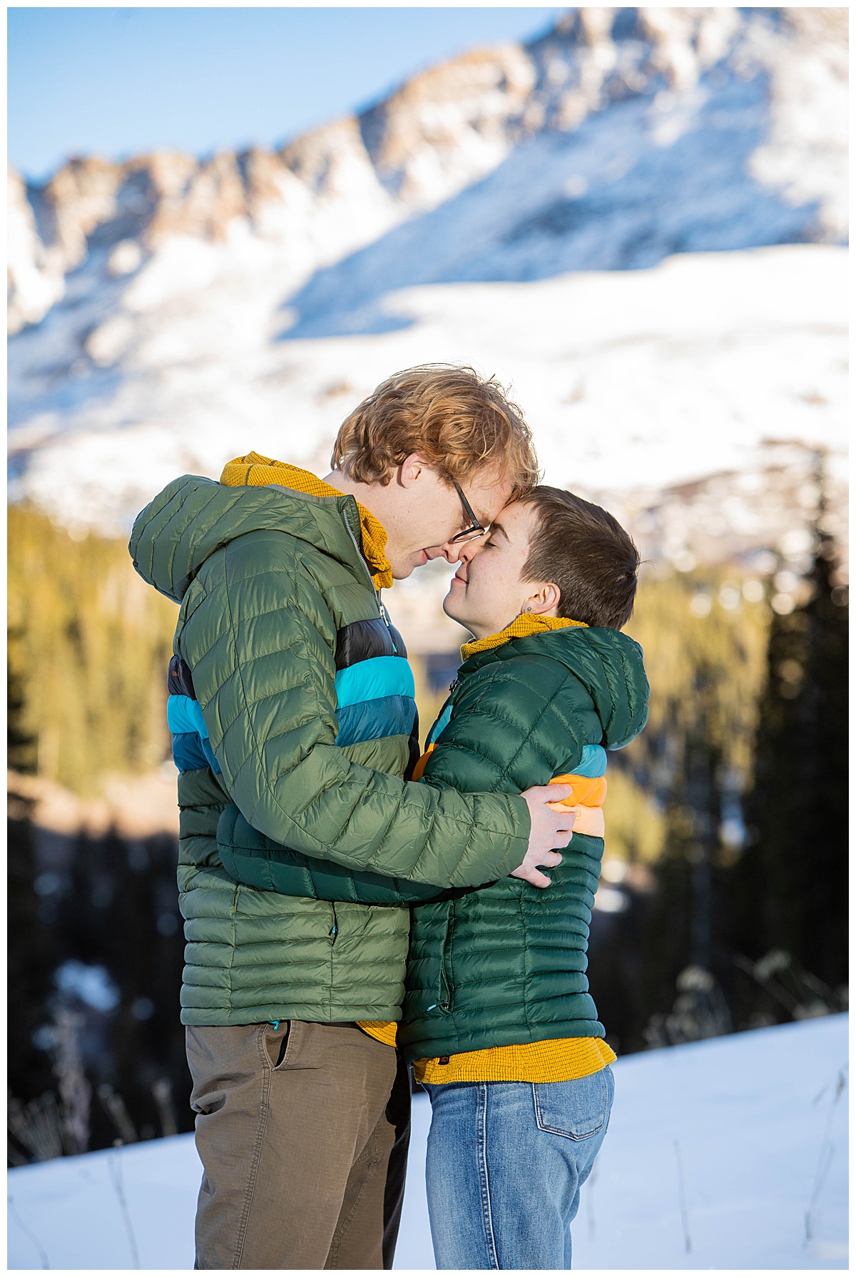 A couple poses in front of snow, mountains, and pine trees.