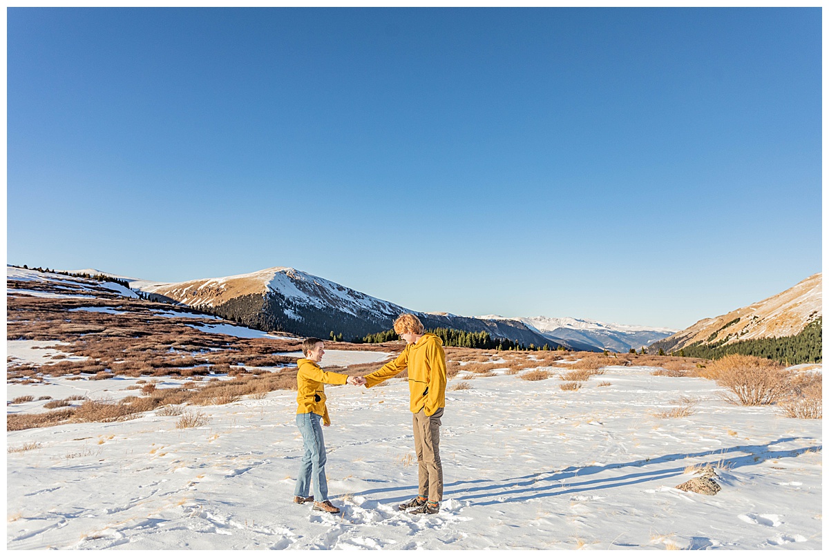 A couple poses in front of snow, mountains, and pine trees.
