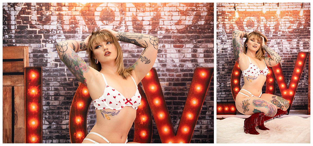 A woman poses for Denver boudoir in white lingerie with little red hearts all over it. Behind her is a background with brick, lights, crates, and large LOVE letters.