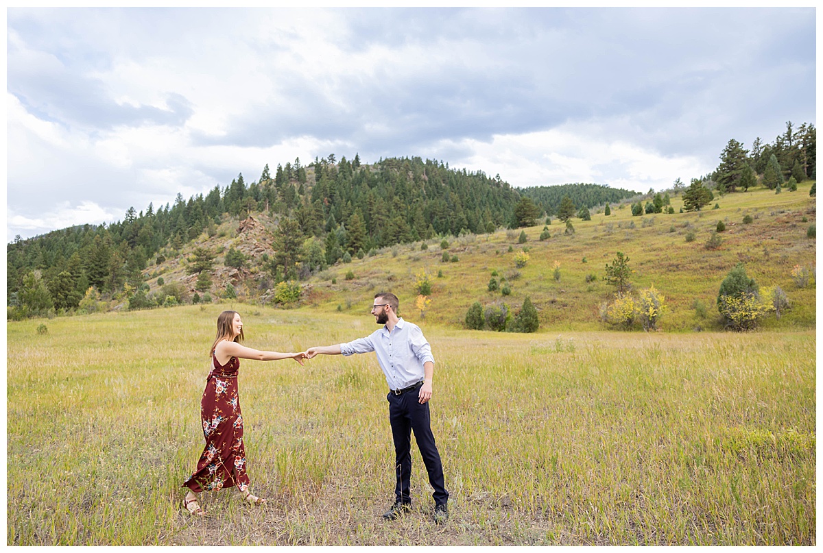 A couple poses dances in a field, posing for a fall engagement session. The woman is wearing a long red dress and the man is wearing a light blue button up and dark navy pants.