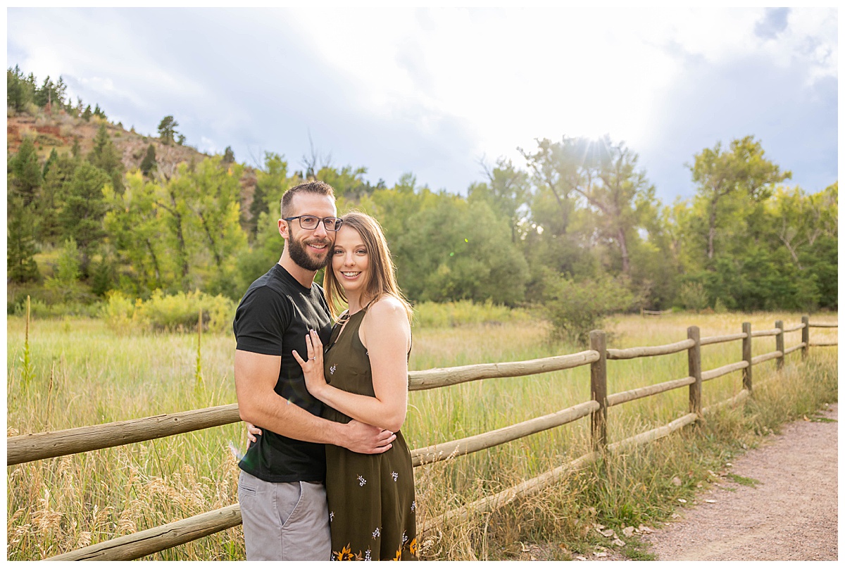 A couple poses on a pathway for a fall engagement session. The woman is wearing a short olive green dress and the man is wearing a black t-shirt and gray shorts.