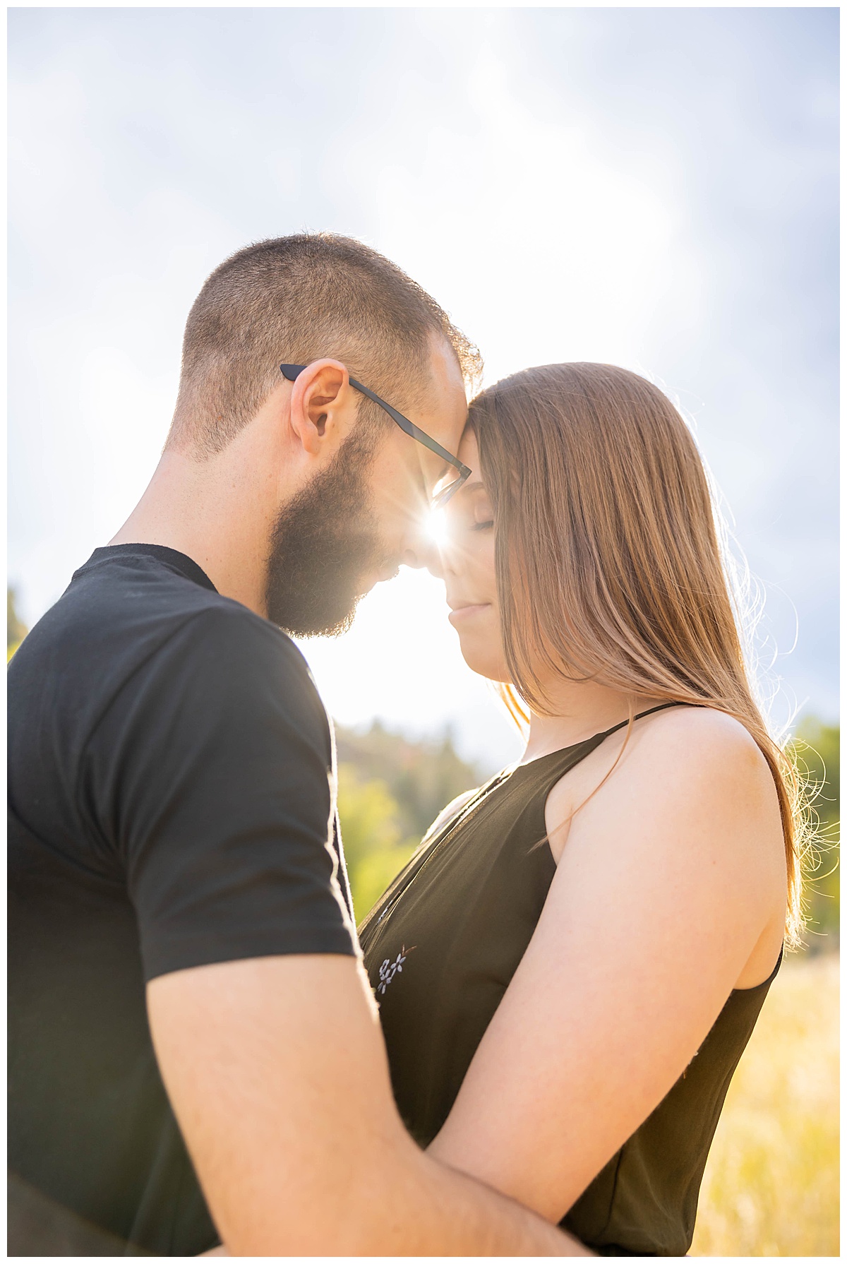 A couple poses with the sun behind them for a fall engagement session. The woman is wearing a short olive green dress and the man is wearing a black t-shirt and gray shorts.