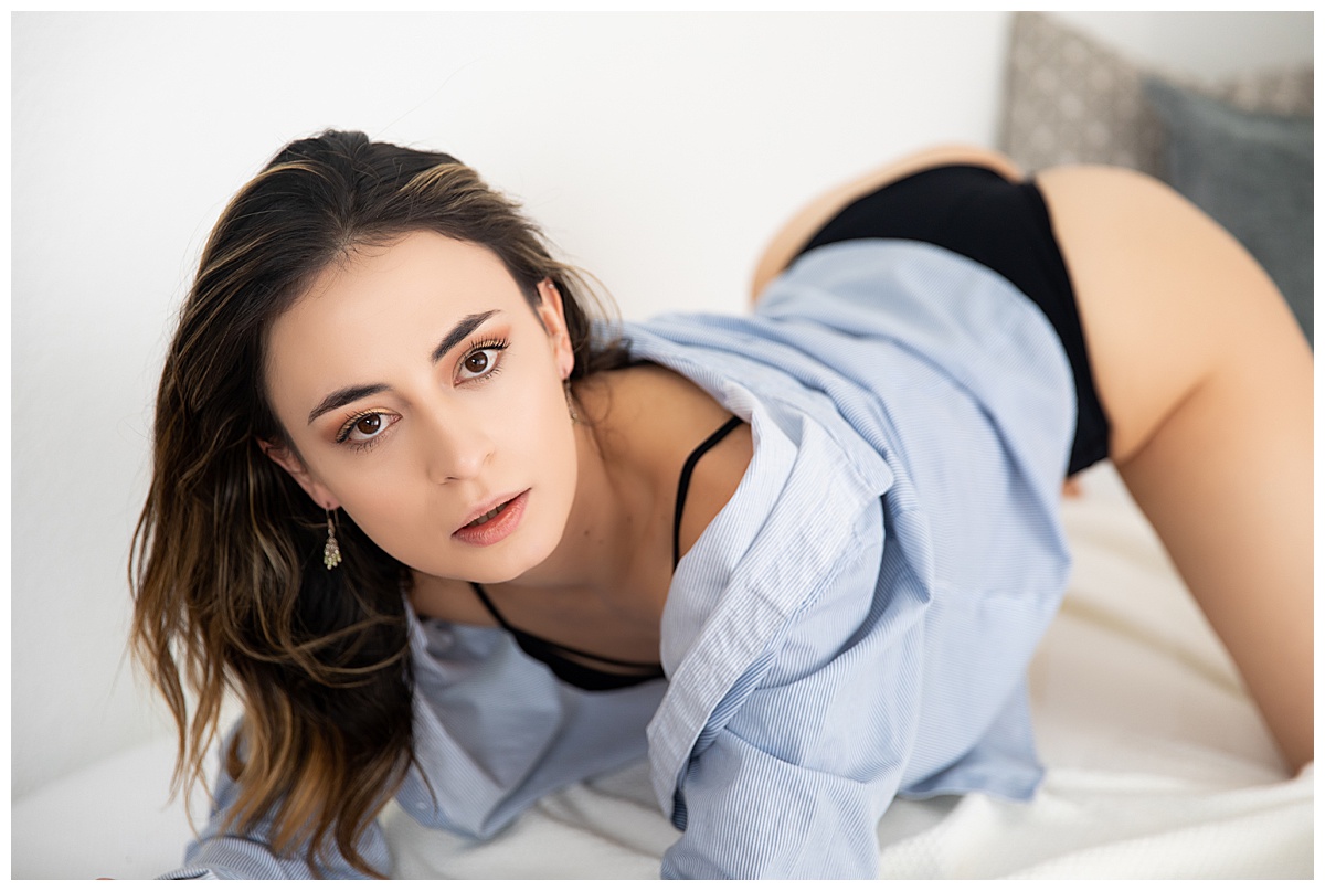 A woman poses on a bed in a blue button down in a spring floral boudoir session