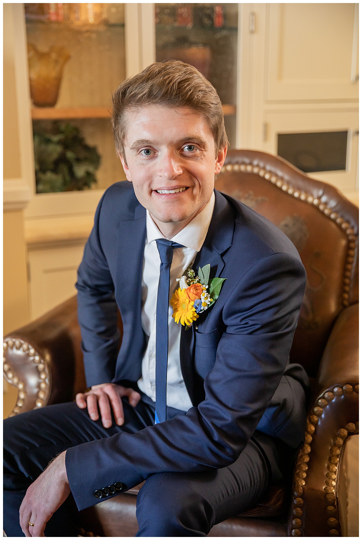 The groom poses in the groom's quarters in his blue suit on a leather chair. 