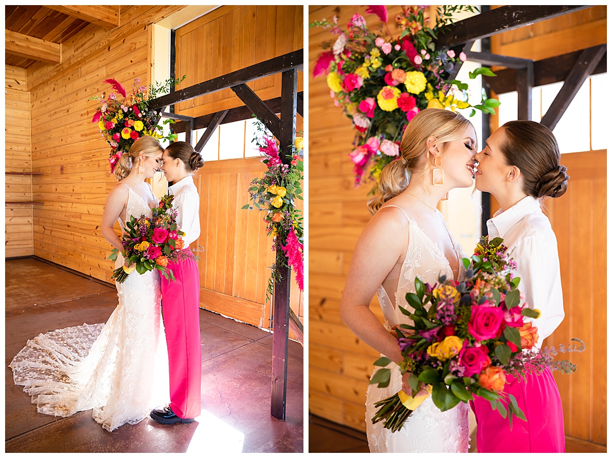 Two brides pose in front of a dark brown wedding arch covered in bright pink and yellow flowers. One bride is blonde, wearing a dress with flowers on it. The other bride is a brunette and is wearing a while button down and hot pink pants.
