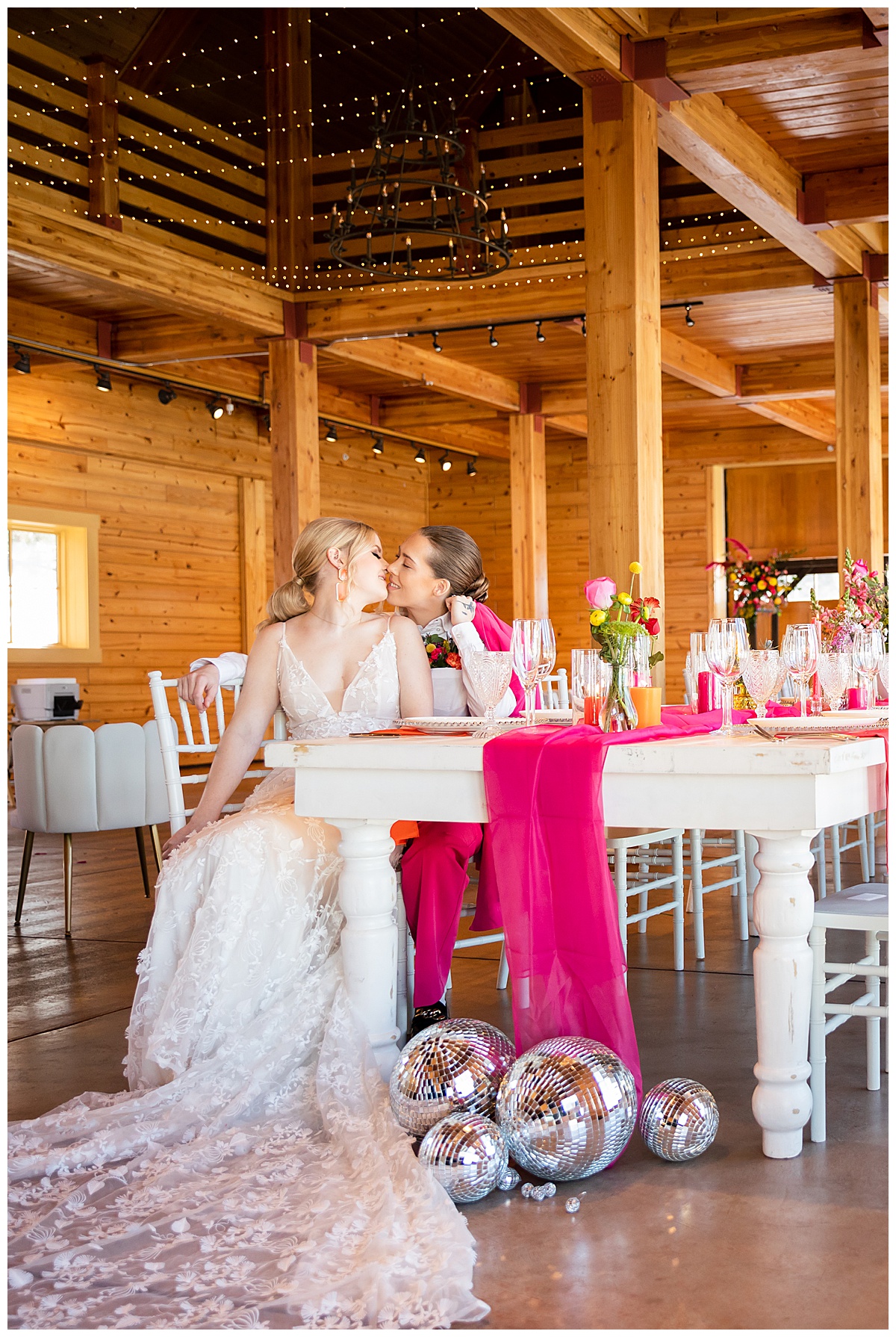 The brides sit at the spring wedding inspiration table with flowers and a hot pink runner. The wood barn is the background, with twinkle lights and a chandelier.