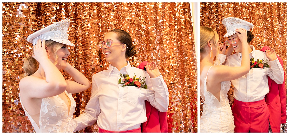 The brides pose in front of a rose gold sparkling background for the photo booth. The bride in the dress is wearing the bedazzled bride hat and the bride in the pants is wearing fun pink sunglasses.