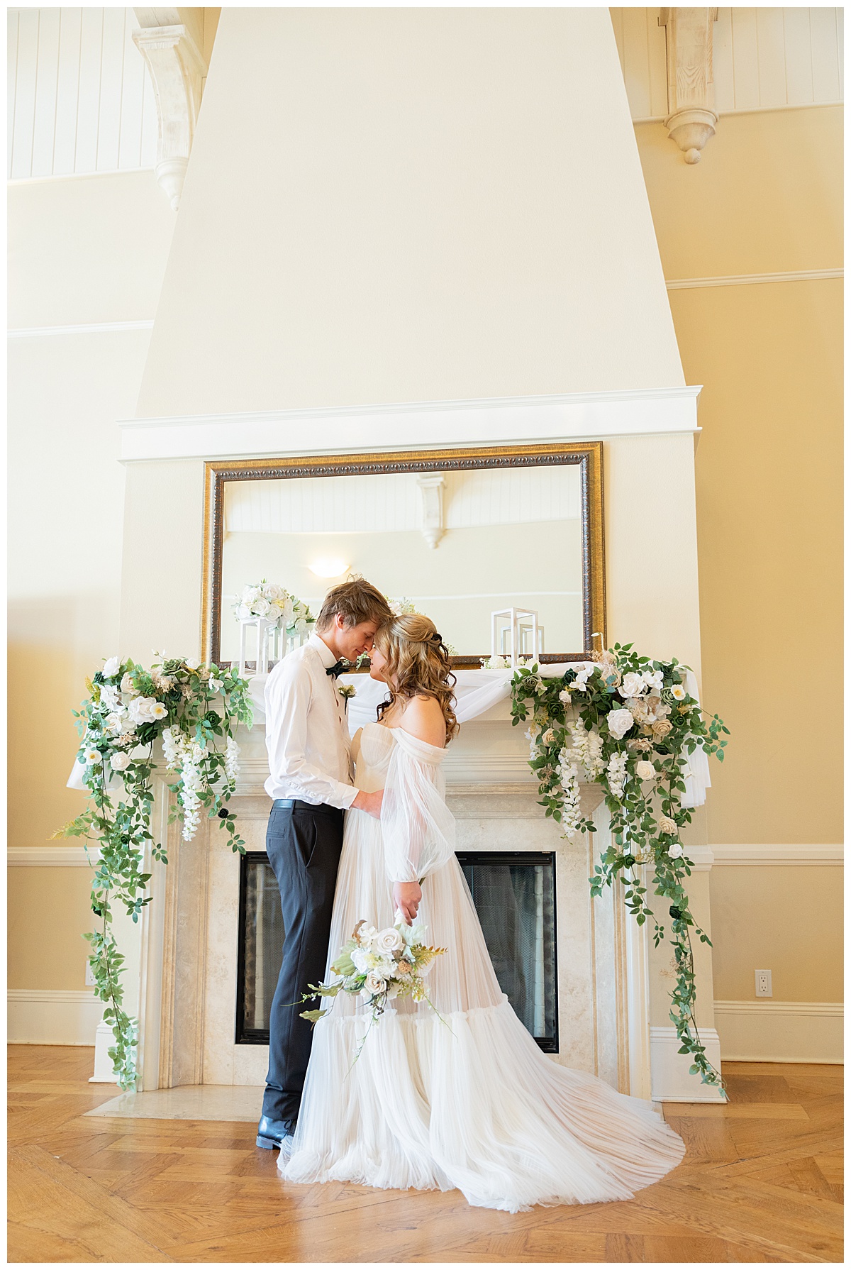 A bride and groom pose in front of a white fireplace with greenery and white flowers all over it for a winter wedding inspiration shoot.