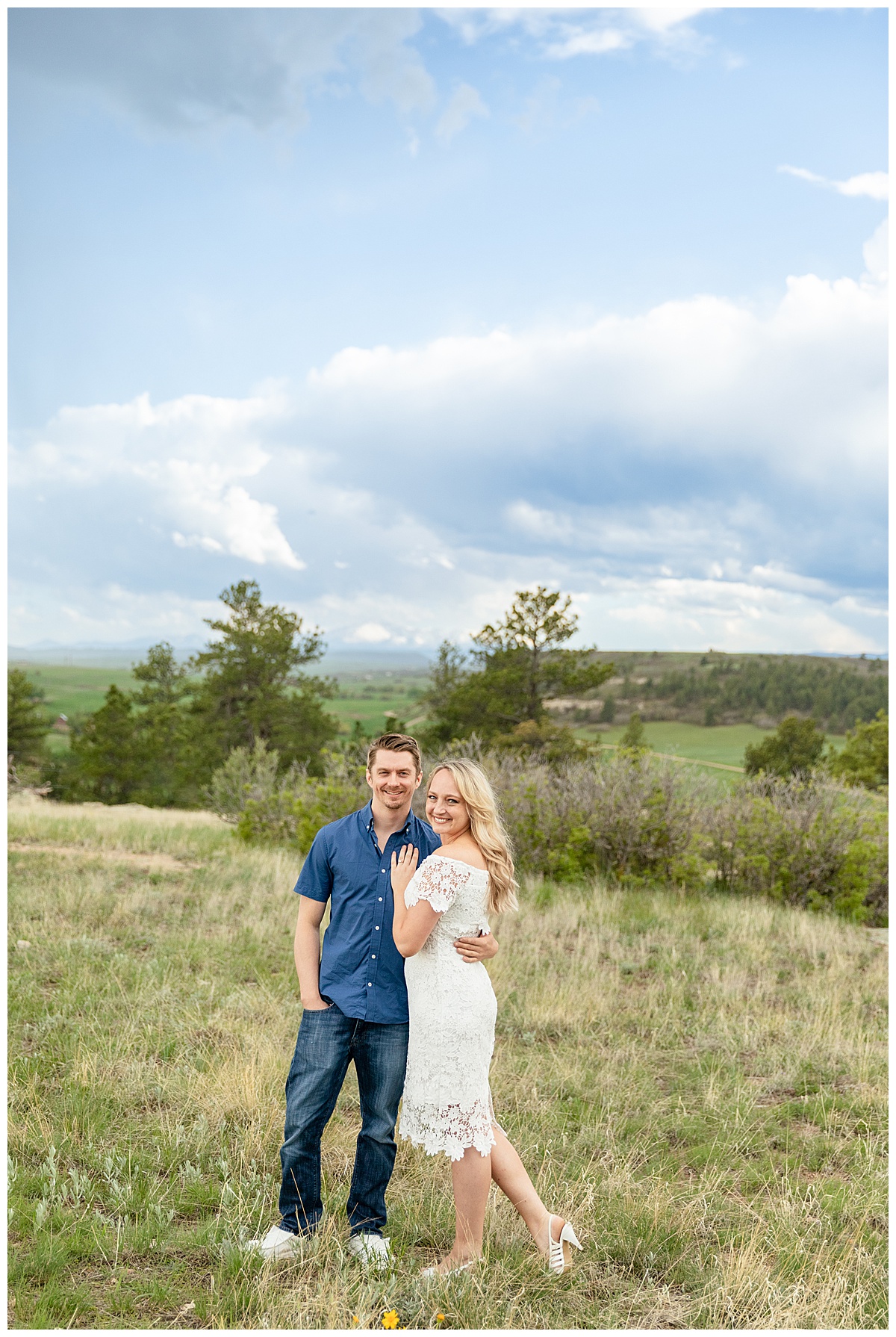A couple poses for their Colorado engagement session in front of trees and mountains. The man is wearing a blue button up shirt. The woman is blond and wearing a white lace dress.