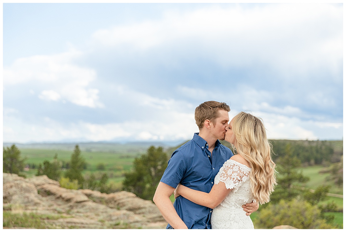 A couple poses for their Colorado engagement session in front of trees and mountains. The man is wearing a blue button up shirt. The woman is blond and wearing a white lace dress.