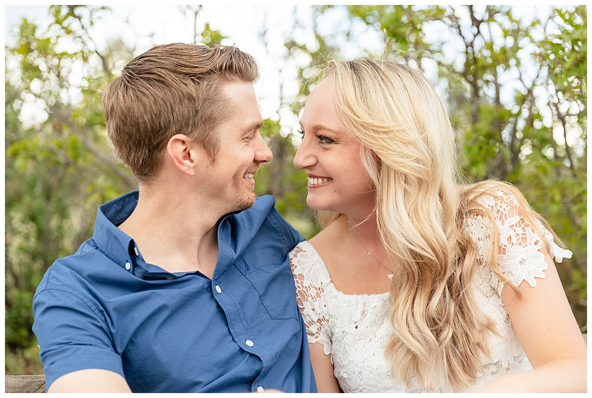 A couple poses for their portraits. The man is wearing a blue button up shirt. The woman is blond and wearing a white lace dress.