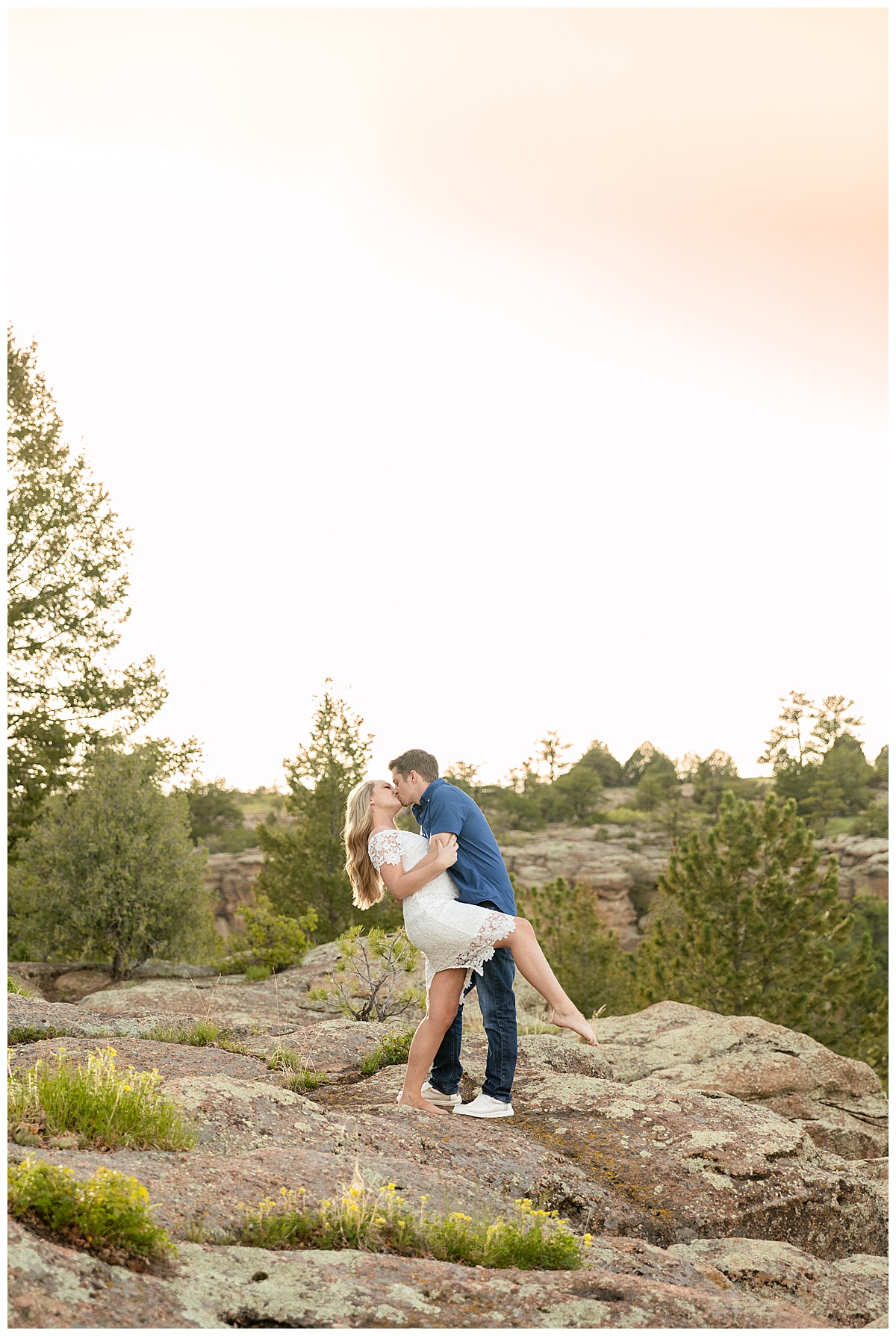 A couple poses for their portraits in front of trees and rocks. The man is wearing a blue button up shirt. The woman is blond and wearing a white lace dress.
