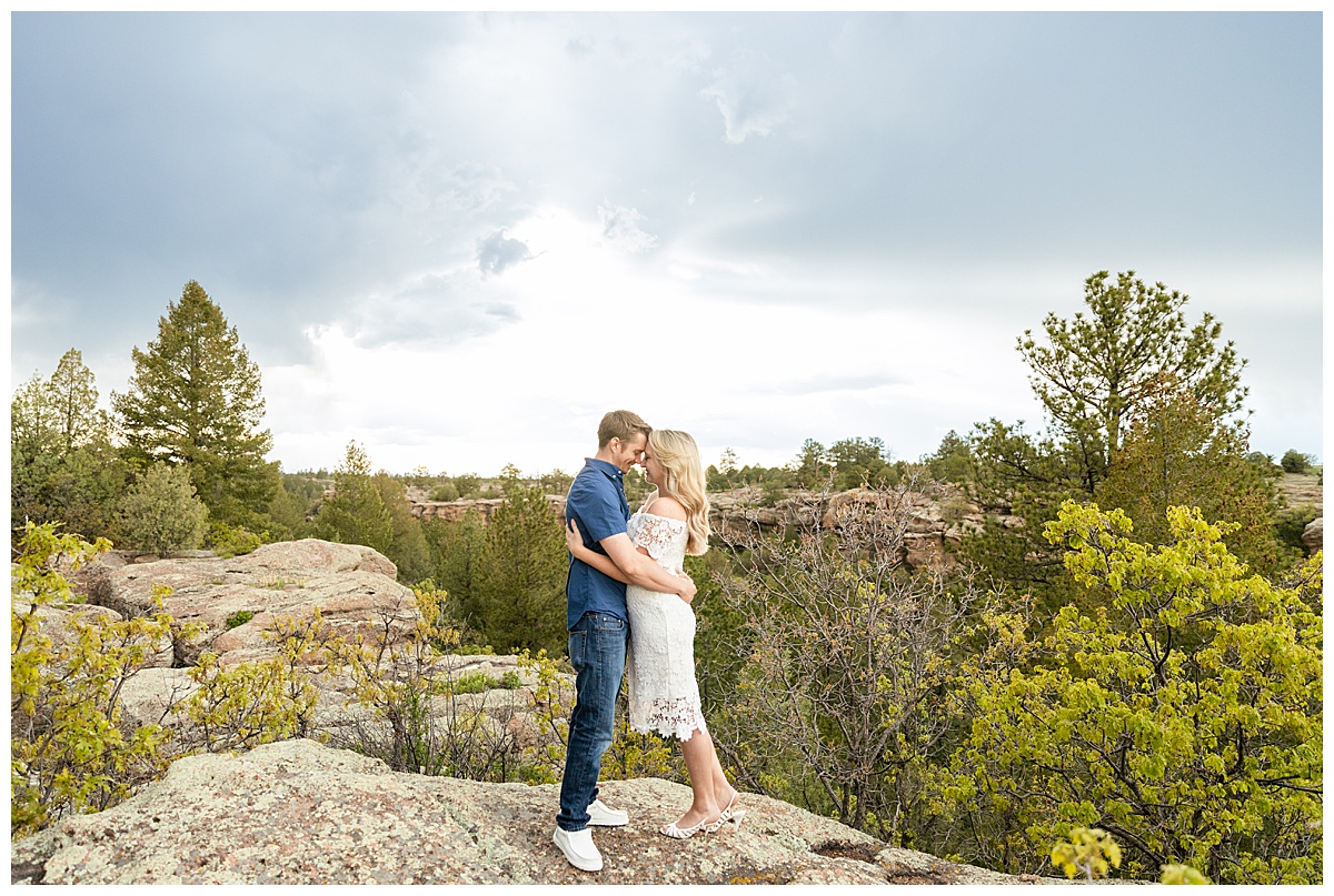 A couple poses for their portraits in front of trees and rocks. The man is wearing a blue button up shirt. The woman is blond and wearing a white lace dress.