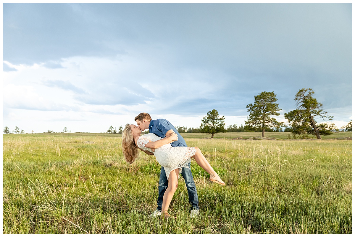 A couple poses for their portraits in a field. The man is wearing a blue button up shirt. The woman is blond and wearing a white lace dress.