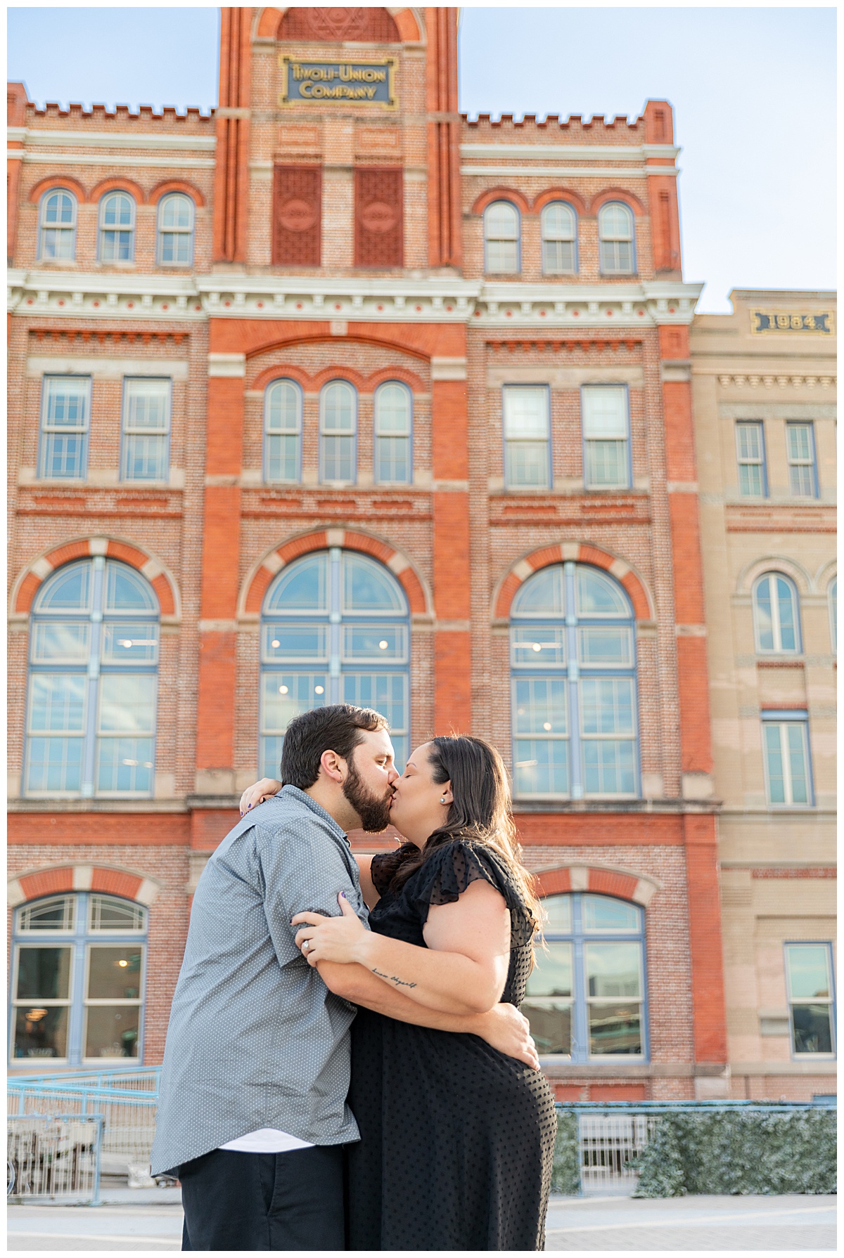 A couple in black and gray pose in front of a large red brick building for their Auraria Campus engagement session