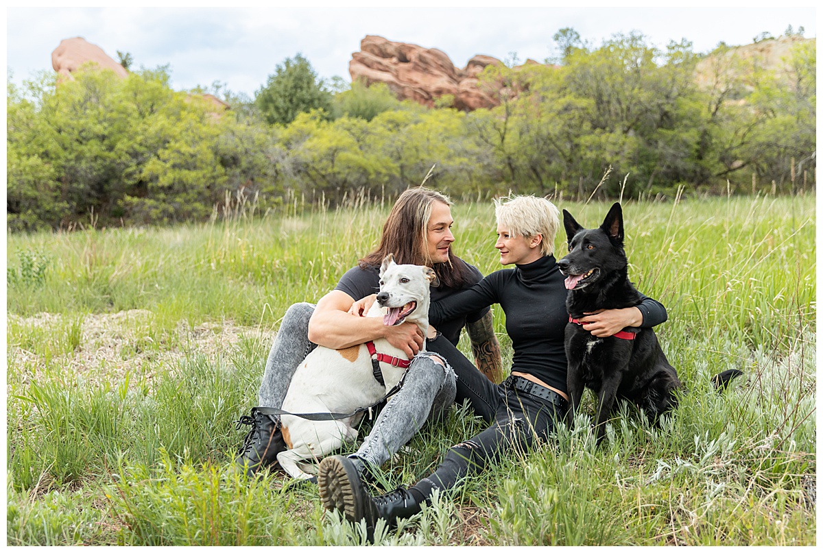 A couple dressed in black pose in front of a field of grass and large red rocks with their dogs for their dog couples session