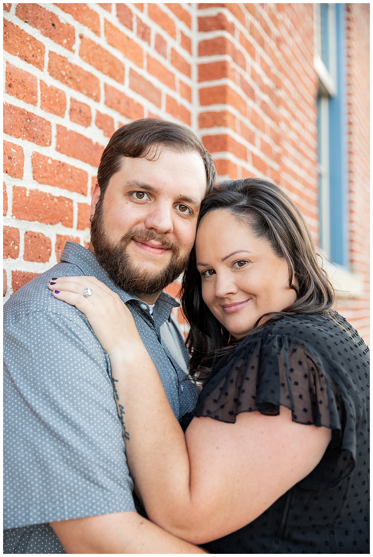 A couple in black and gray pose in front of a large red brick building for their Auraria Campus engagement session