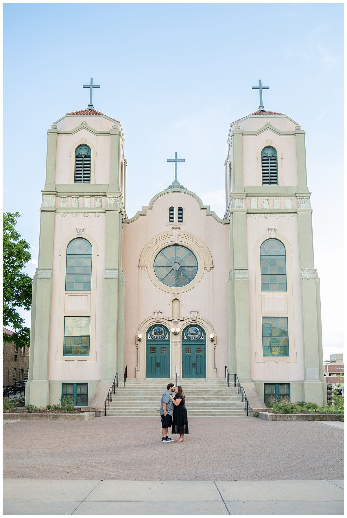 A couple in black and gray pose in front of an old pink and green church