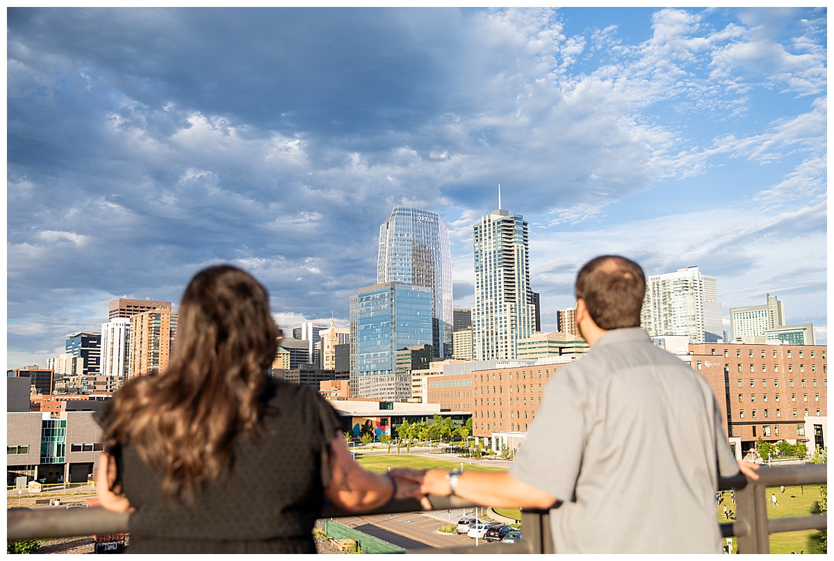 A couple in black and gray pose on top of a parking garage overlooking the city skyline of Denver, CO