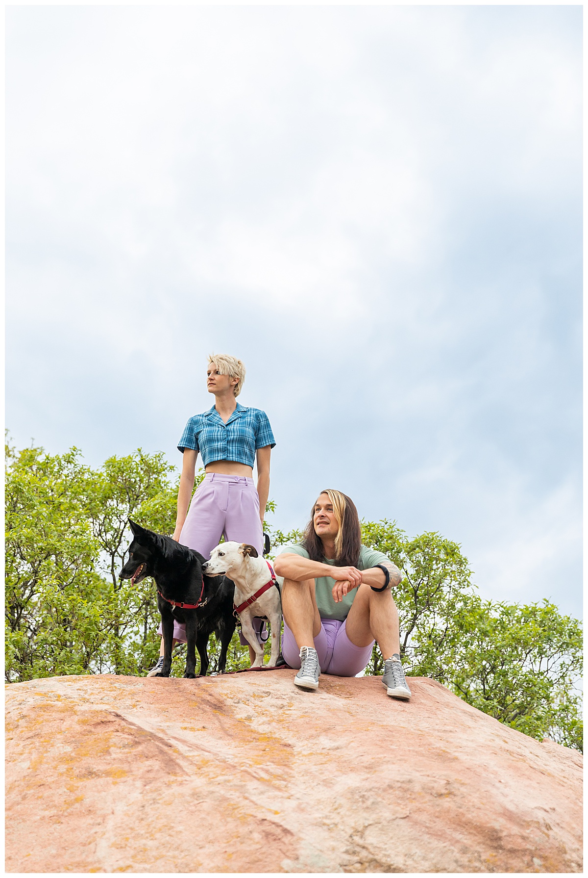 A couple dressed in purple, blue, and green pose on top of a large red rock with their dogs at their dog couples session