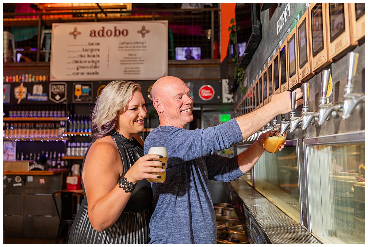A woman with blonde hair dark silver dress and a bald man in a blue sweater pose with beers at their brewery engagement session