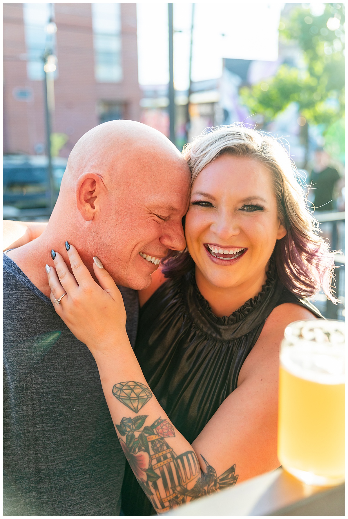 A woman with blonde hair dark silver dress and a bald man in a blue sweater pose with beers at their brewery engagement session