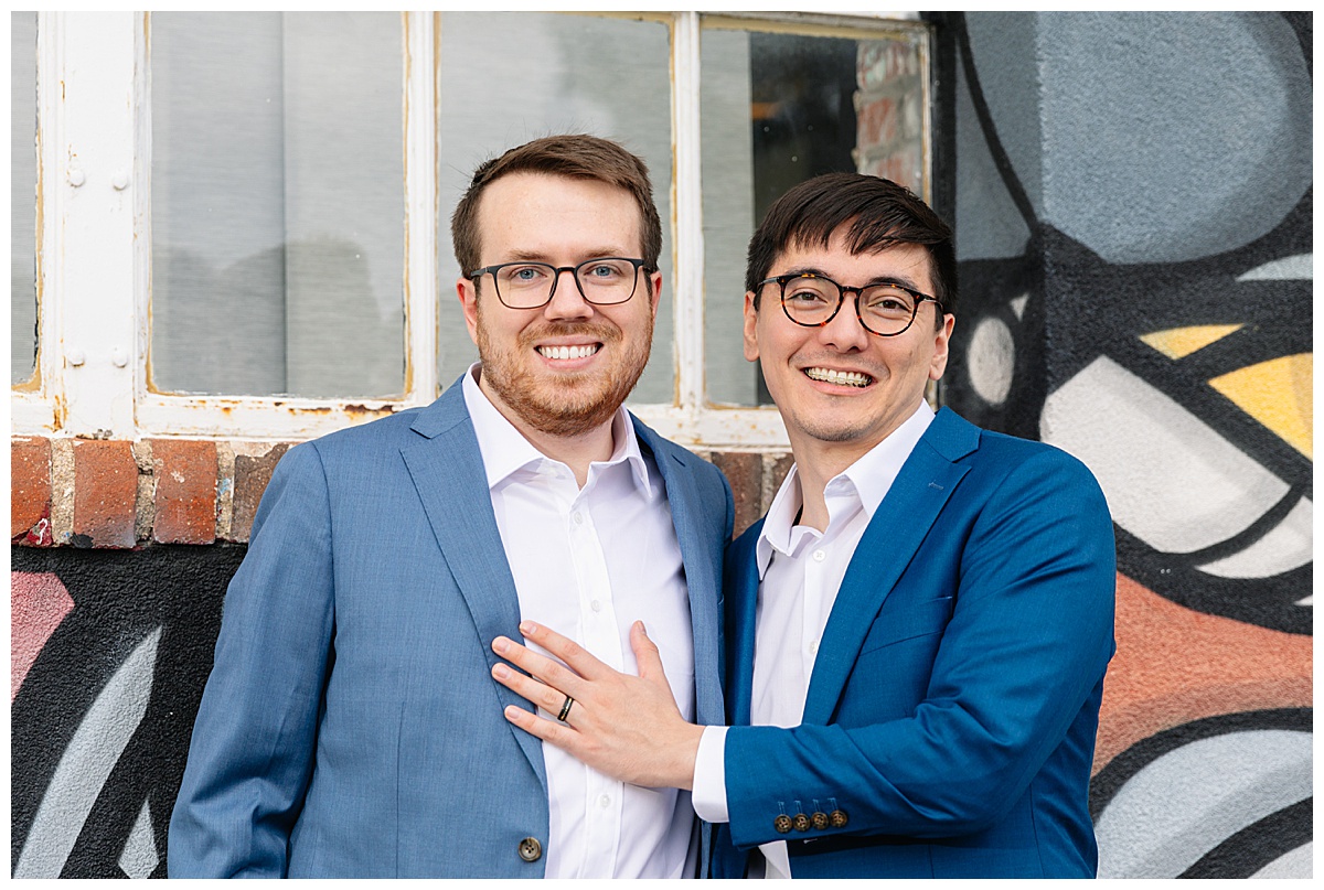 Two men pose in front of a graffitied wall dressed in blue suits for their RiNo engagement session