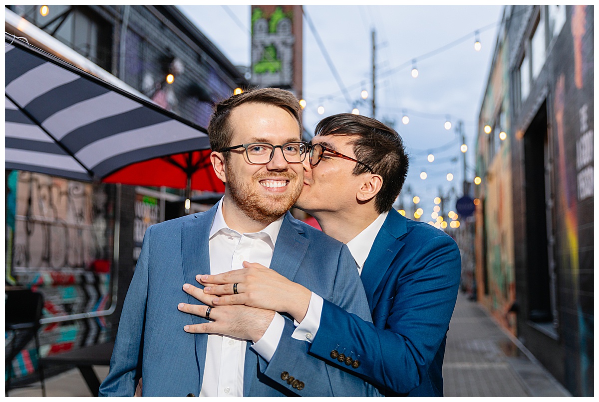 Two men pose in front of an alleyway with twinkle lights dressed in blue suits for their RiNo engagement session