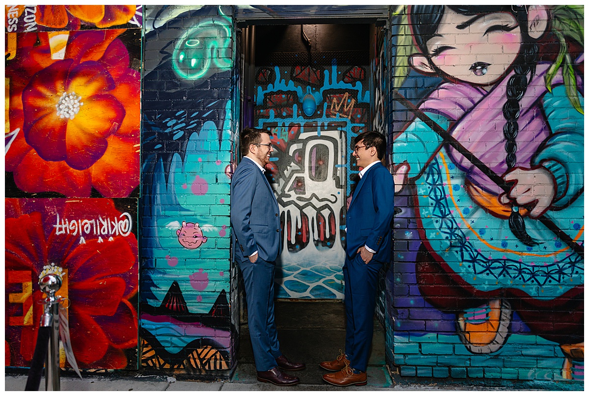 Two men pose in front of a graffitied wall dressed in blue suits