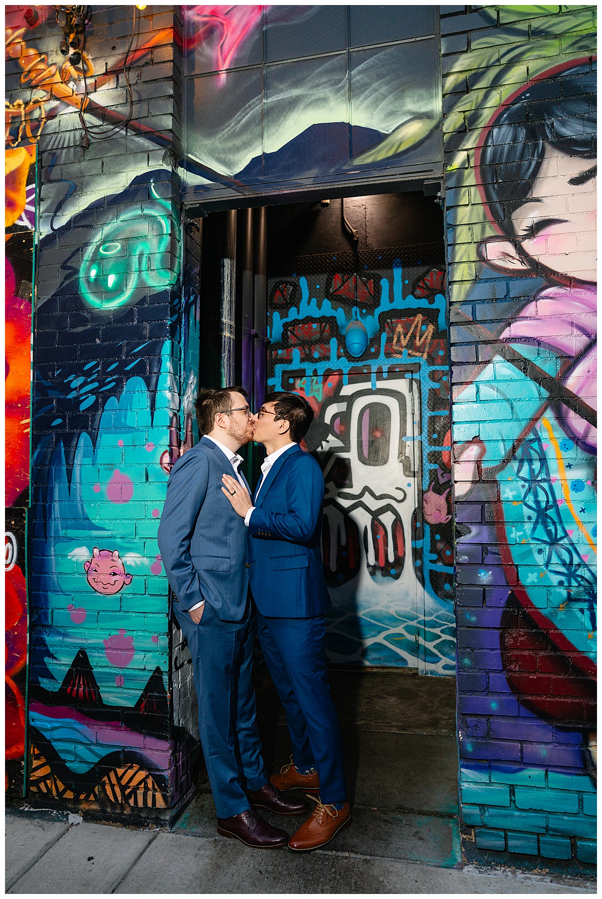 Two men pose in front of a graffitied wall dressed in blue suits