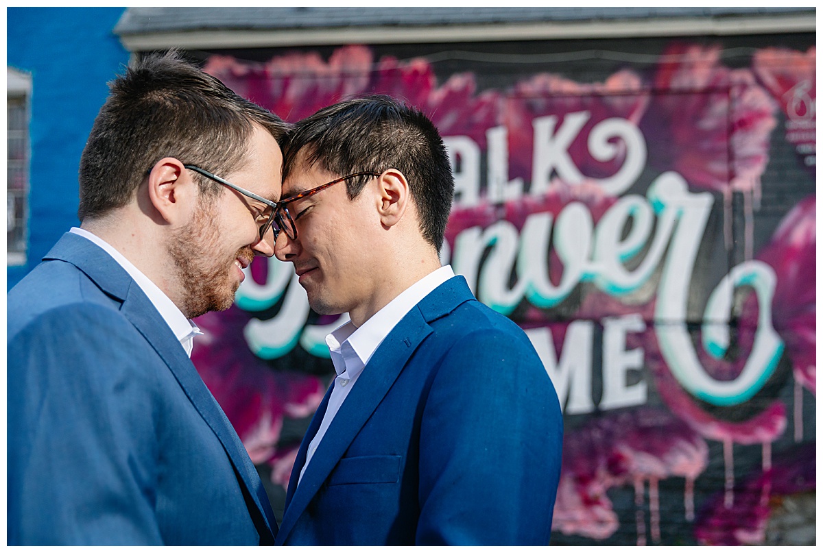 Two men pose in front of a graffitied wall dressed in blue suits for their RiNo engagement session