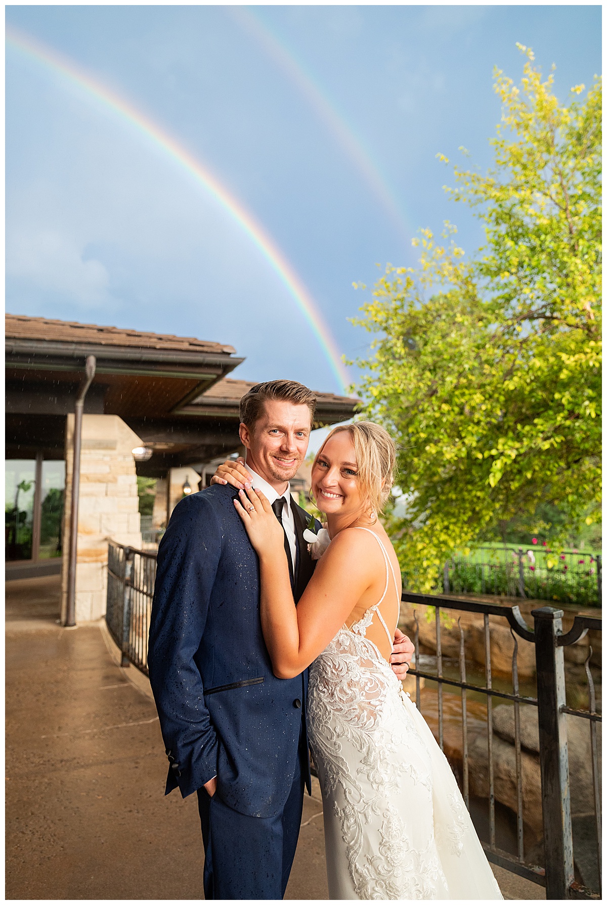 A bride in a white dress and a groom in a navy suit pose in the rain in front of a double rainbow on their wedding day