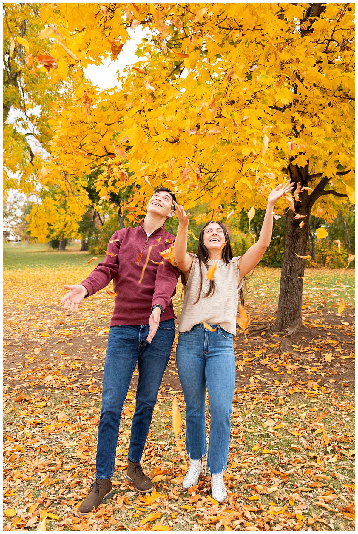 A couple poses in front of a bright yellow tree. The man is wearing a red sweater and blue jeans, the woman is wearing a sleeveless cream sweater and blue jeans.