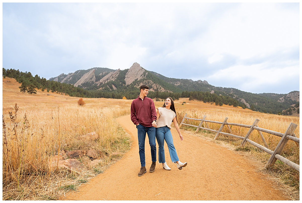A couple poses in front of the Boulder flatirons. The man is wearing a red sweater and blue jeans, the woman is wearing a sleeveless cream sweater and blue jeans.