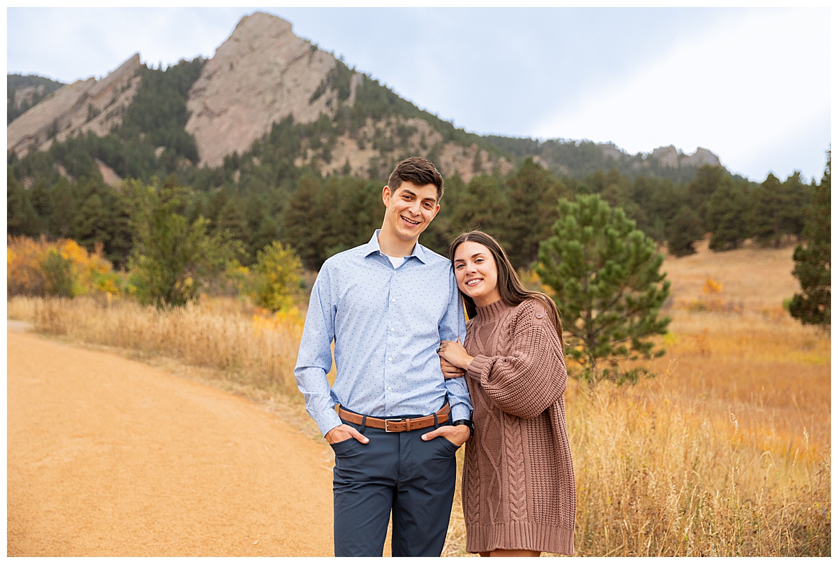 A couple poses in front of the Boulder flatirons for their Chautauqua Park engagement session. The man is wearing a blue button down and navy dress pants, the woman is wearing a mauve sweater dress.
