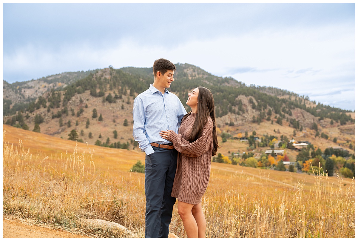 A couple poses overlooking Boulder, CO. The man is wearing a blue button down and navy dress pants, the woman is wearing a mauve sweater dress.