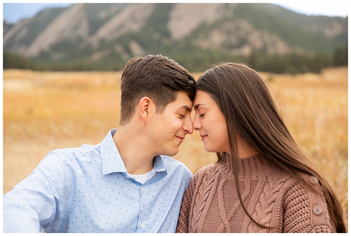 A couple poses in front of the Boulder flatirons for their Chautauqua Park engagement session. The man is wearing a blue button down and navy dress pants, the woman is wearing a mauve sweater dress.