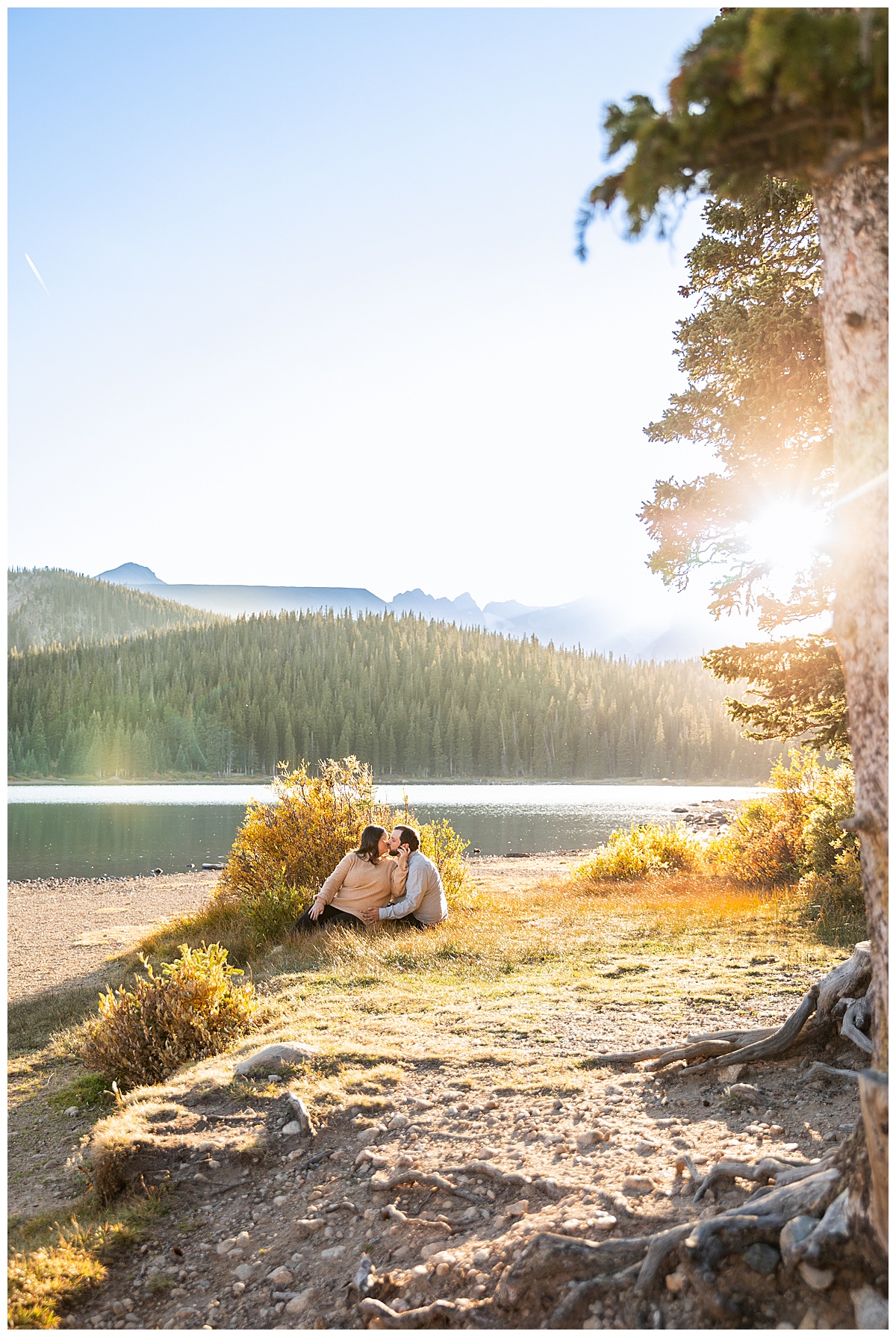 A couples poses for their fall lake engagement session in front of yellow bushes, a lake, and mountains. The man is wearing a gray long sleeve button down shirt and the woman is wearing a tan sweater