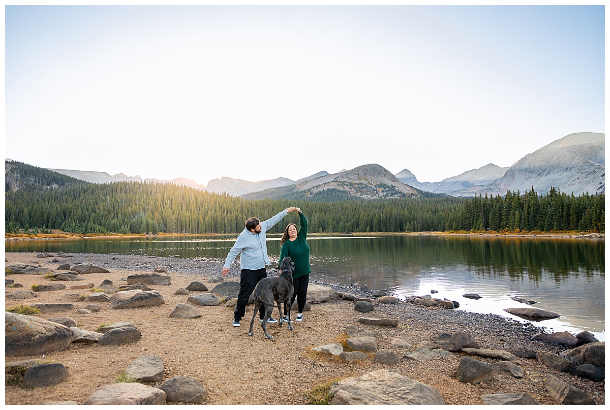 A couples poses for their fall lake engagement session with their gray great dane in front of a lake and mountains. The man is wearing a gray long sleeve button down shirt and the woman is wearing a dark green sweater