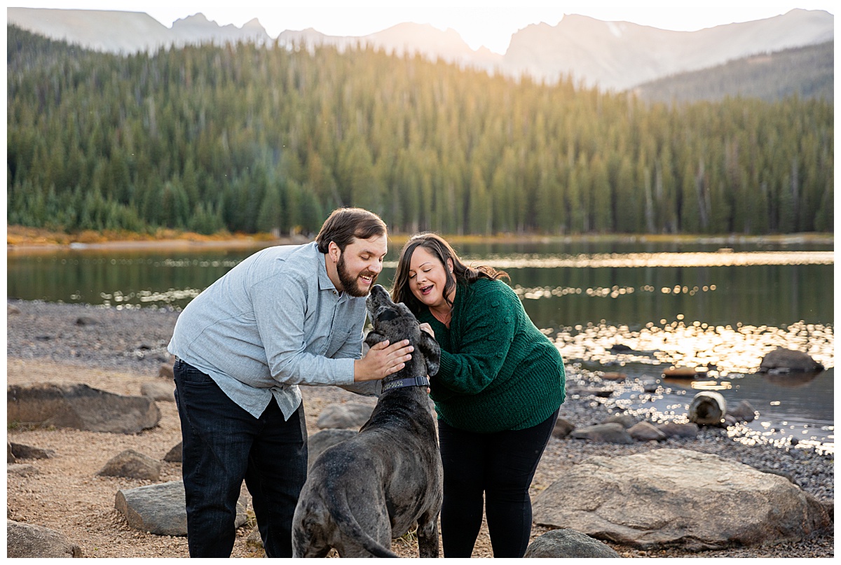 A couple cuddles with their great dane in front of an alpine lake, pine trees, and mountains. The man wears a gray button down and jeans. The woman wears a green sweater and leggings.