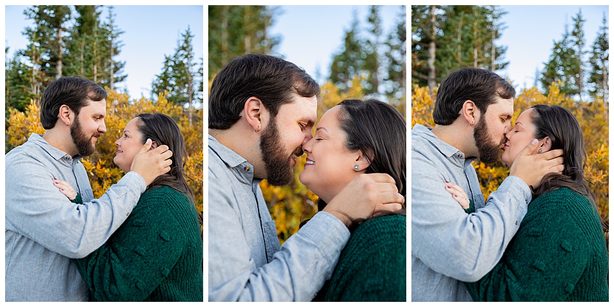 A couples poses in front of yellow bushes. The man is wearing a gray long sleeve button down shirt and the woman is wearing a dark green sweater