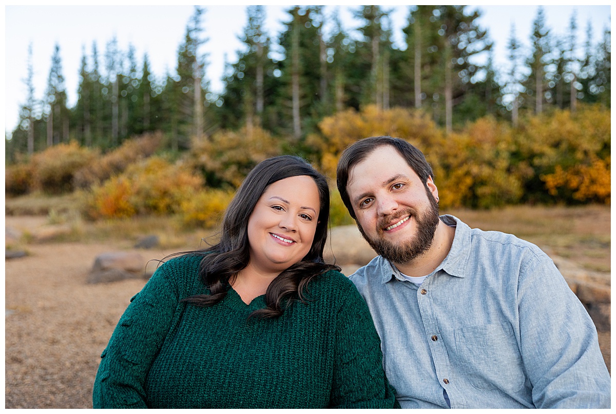 A couples poses in front of yellow bushes and pine trees. The man is wearing a gray long sleeve button down shirt and the woman is wearing a dark green sweater