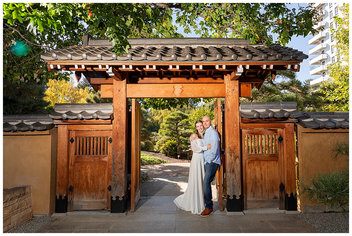 A couple poses in a wooden gateway during their botanic gardens engagement session. The blonde woman is wearing a white long sleeve dress and the bald man is wearing a blue plaid shirt and jeans.
