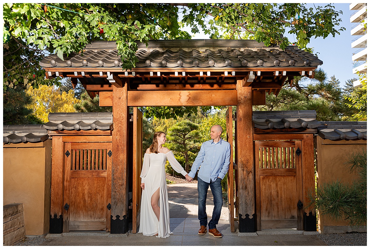 A couple poses in a wooden gateway during their botanic gardens engagement session. The blonde woman is wearing a white long sleeve dress and the bald man is wearing a blue plaid shirt and jeans.
