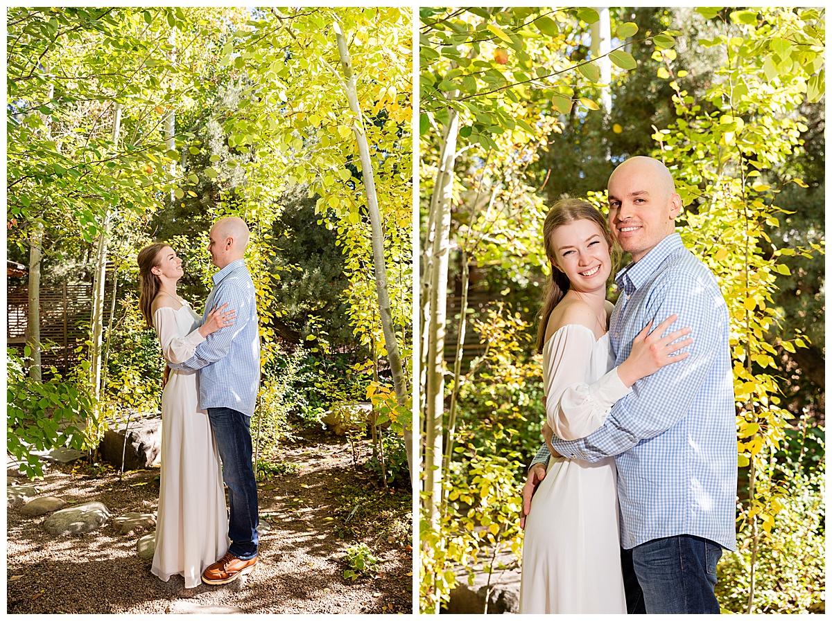 A couple poses in front of trees. The blonde woman is wearing a white long sleeve dress and the bald man is wearing a blue plaid shirt and jeans.