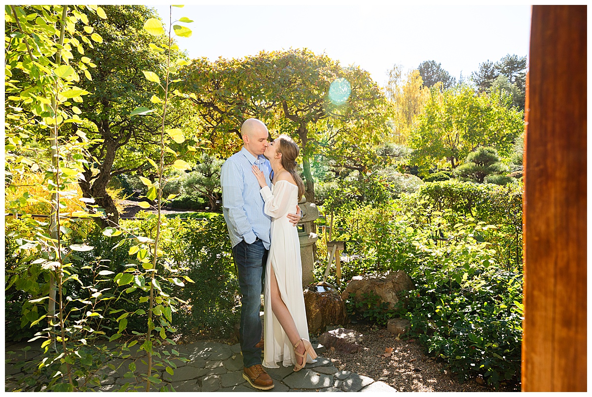 A couple poses in front of trees and a stone lantern. The blonde woman is wearing a white long sleeve dress and the bald man is wearing a blue plaid shirt and jeans.