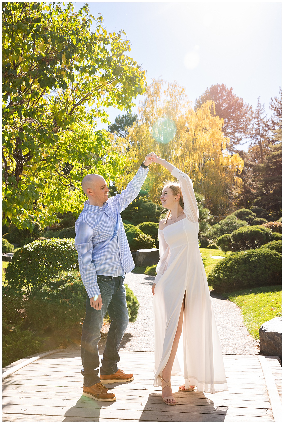 A couple dances on a pathway in front of trees and bushes during their engagement session. The man is spinning the woman. The man is wearing a blue checkered shirt and jeans. The woman is wearing a long white dress with long sleeves.