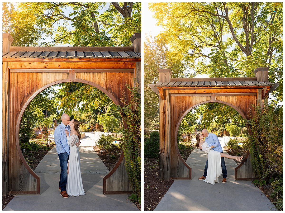 A couple poses in a wooden archway during their botanic gardens engagement session. The blonde woman is wearing a white long sleeve dress and the bald man is wearing a blue plaid shirt and jeans.