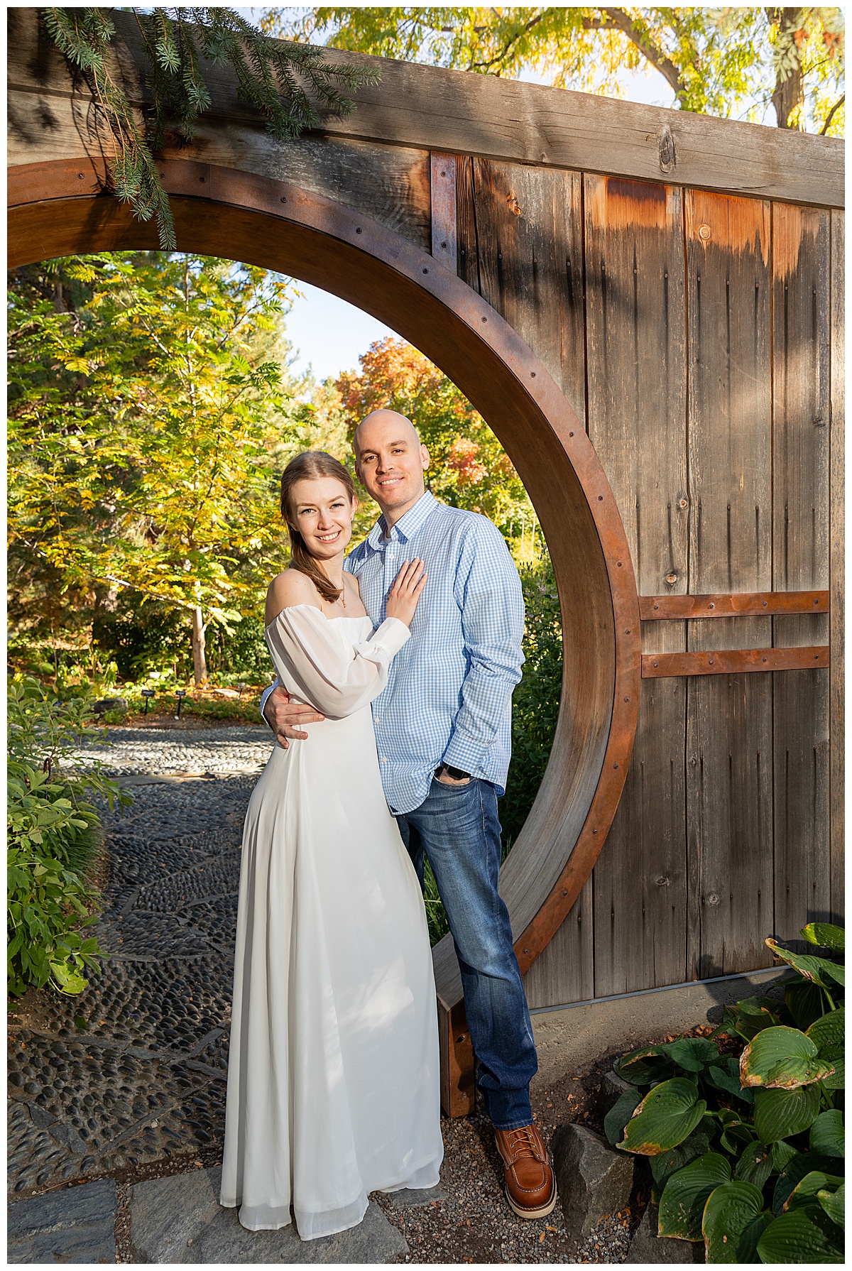 A couple poses in a wooden archway during their botanic gardens engagement session. The blonde woman is wearing a white long sleeve dress and the bald man is wearing a blue plaid shirt and jeans.