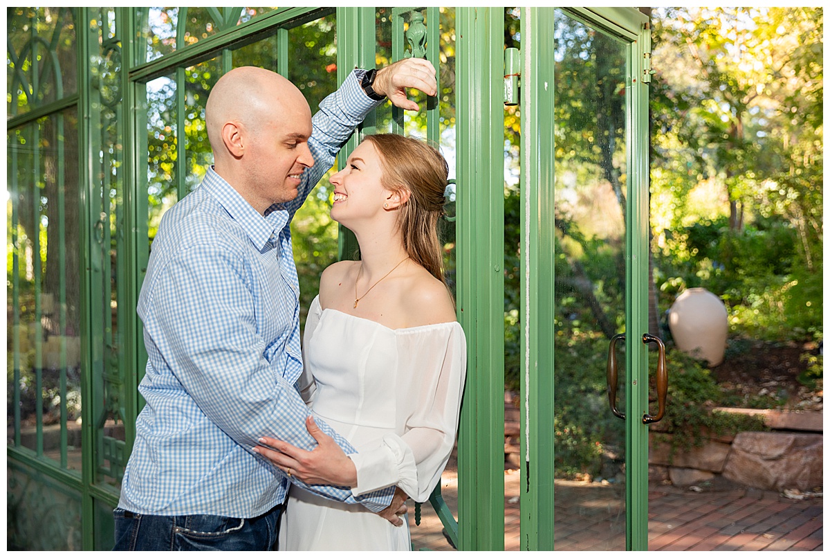 A couple poses inside of a green glass solarium. The blonde woman is wearing a white long sleeve dress and the bald man is wearing a blue plaid shirt and jeans.